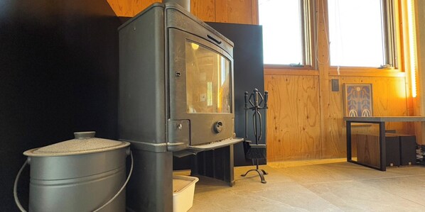 A wood-burning stove, a trash bin, and a window with curtains.