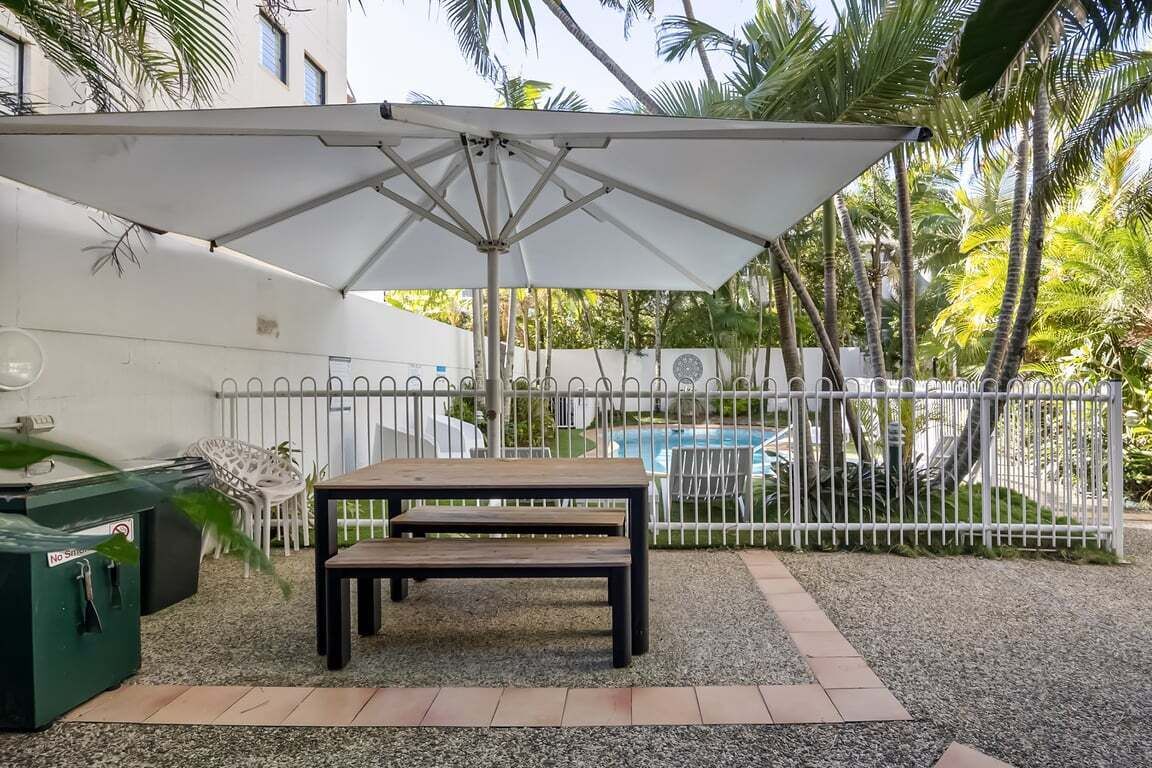 An outdoor patio with a table and umbrella, surrounded by a white fence, palm trees, and a pool.