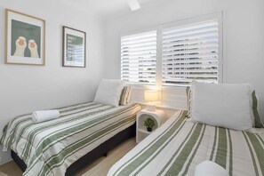 A bedroom with two beds, white bedding, a bedside table with a lamp, and a window with blinds.