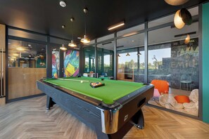 Uma mesa de bilhar em um quarto moderno com grandes janelas, piso de madeira e iluminação pendente.