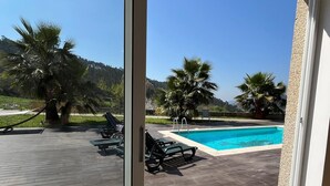 Udsigt gennem en skydedør til et poolområde med træterrasse, liggestole og palmer.