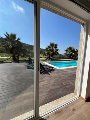 A view through a sliding glass door to a pool area with a wooden deck, lounge chairs, and palm trees. - Charming room 4 metres from the pool, guaranteed relaxation! (GODINHAÇOS)