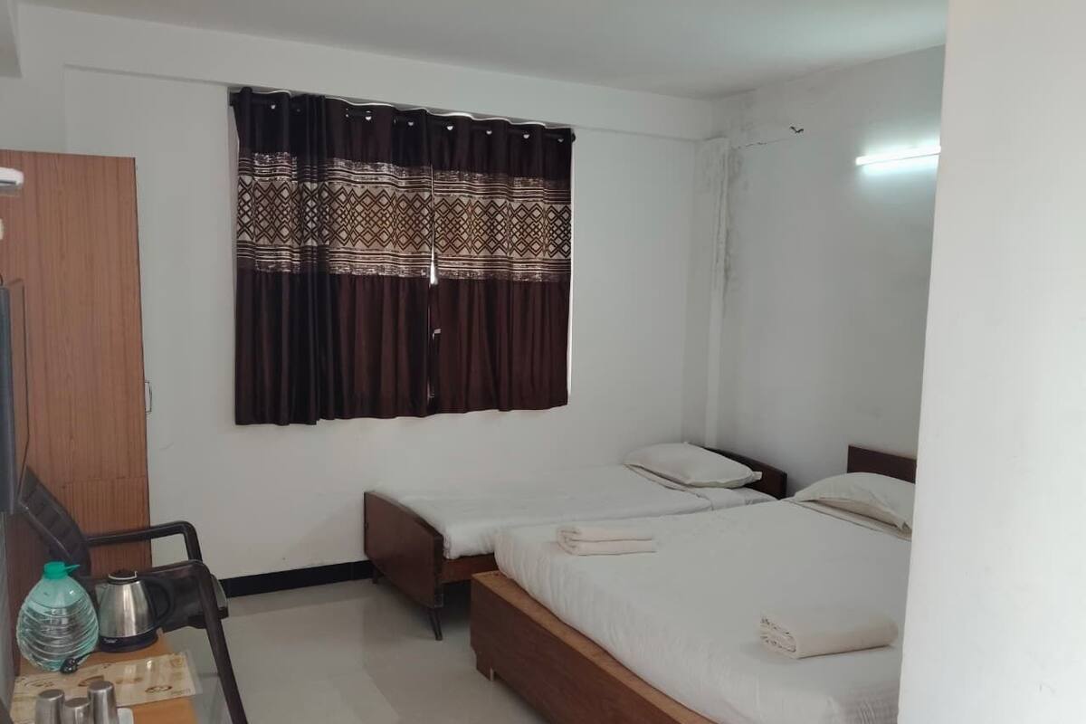 Habitación de hotel con dos camas individuales, un escritorio de madera con una silla, una mesita con una botella de agua y una tetera, y una cortina estampada.