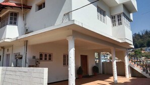 A white multi-story building with a covered patio area, potted plants, and a staircase leading to an upper level. - Woodcock Cottage (Ooty)