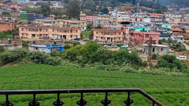 A residential area with houses, a green field, and a metal railing. - Woodcock Cottage (Ooty)