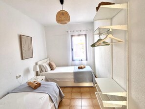 Ett minimalistiskt sovrum med två sängar, en hängande lampa, ett fönster med gardiner och en väggmonterad garderob.