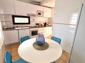 Et kompakt køkken med hvide skabe, en mikroovn, en brødrister og en vask. Der er et rundt bord med en blå vase og to blå stole.