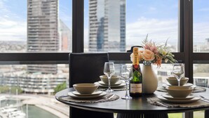 A dining table set with bowls, wine glasses, and a bottle of champagne, with a cityscape view through large windows. - Docklands Marina Waterfront Luxury with 180° Views Pool, Gym & Carpark (Docklands)