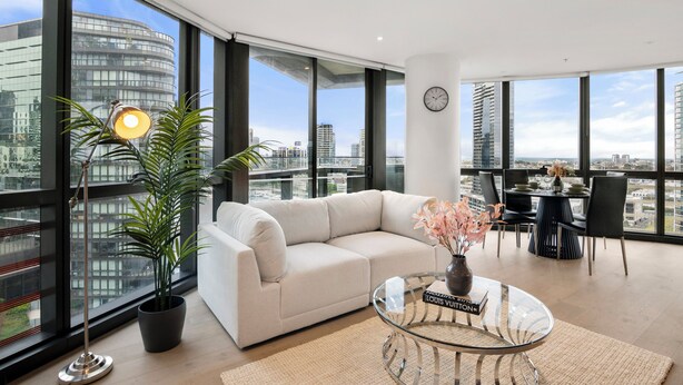 A modern living room with a sofa, a dining table, and a large window with a city view. - Docklands Marina Waterfront Luxury with 180° Views Pool, Gym & Carpark (Docklands)
