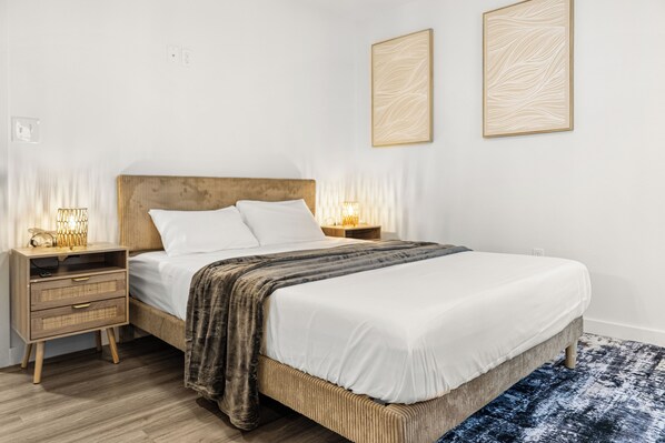 A bedroom with a wooden headboard, a white bedspread, two bedside lamps, and a blue patterned rug. - Modern & Cozy 1 Bedroom Apartment in Baton Rouge (Baton Rouge)