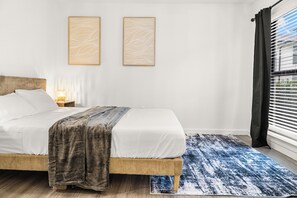 Un lit avec une tête de lit en bois, un tapis à motifs bleus, deux œuvres d’art accrochées au mur et une fenêtre avec des stores.