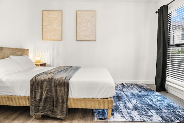 Ein Bett mit hölzernem Kopfteil, ein Teppich mit blauem Muster, zwei an der Wand befestigte Kunstwerke und ein Fenster mit Jalousien.