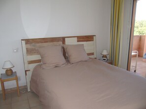 A bedroom with a wooden headboard, two pillows, a bedside table with a lamp, and a window with curtains. - Res. Suarella, a beautiful, fully equipped two-bedroom apartment with breathtaking views of the Gulf of Saint-Florent! (Saint-Florent)