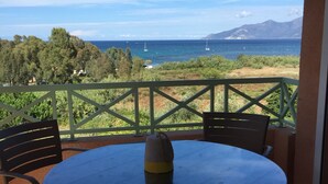 Mesa redonda con sillas en un balcón con vistas a la costa, árboles y una montaña a lo lejos.