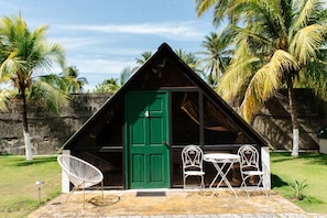 Una pequeña estructura con techo triangular, una puerta verde, dos sillas blancas y una mesa afuera.