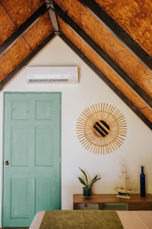 A room with a wooden ceiling, a green door, a wall-mounted air conditioner, a decorative sunburst mirror, a small plant, and a model sailboat on a wooden shelf.