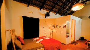 A single bed with a red blanket, a standing fan, a small table with a lamp, and a door leading to a bathroom.