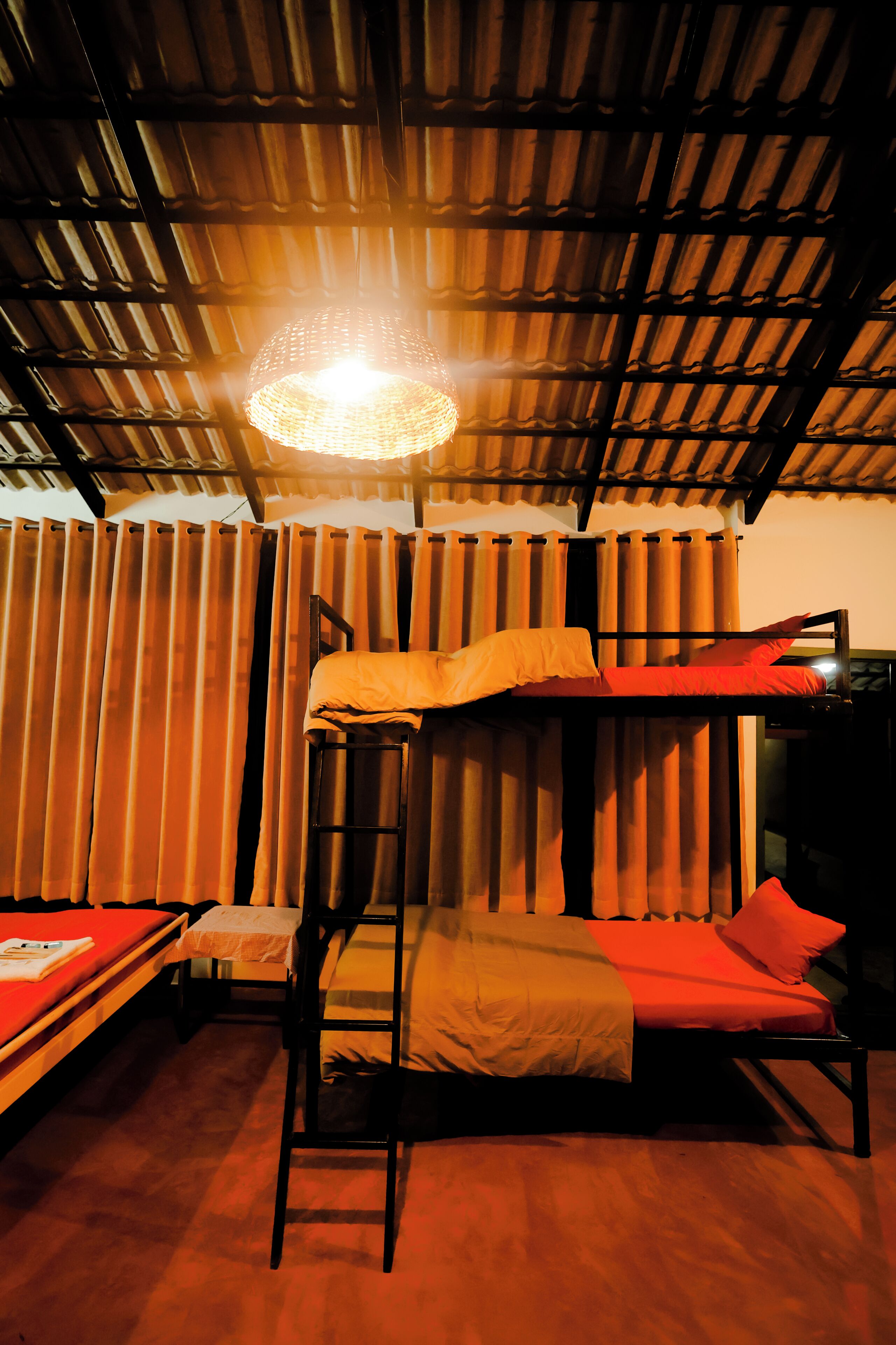 A bunk bed with a ladder, a table with a book, and a hanging light fixture in a room with wooden slats on the ceiling and curtains.