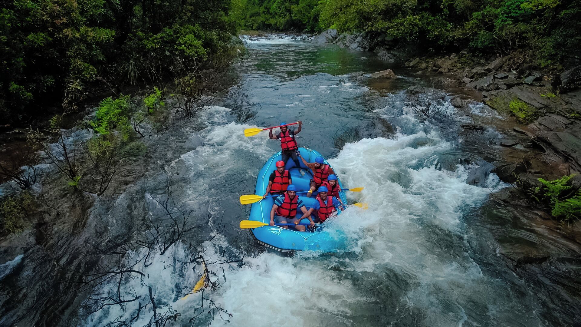 A blue inflatable raft with passengers paddling through a river surrounded by rocky banks and dense greenery.