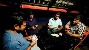 Four people playing cards at a table in a dimly lit room with a black table and red chairs.