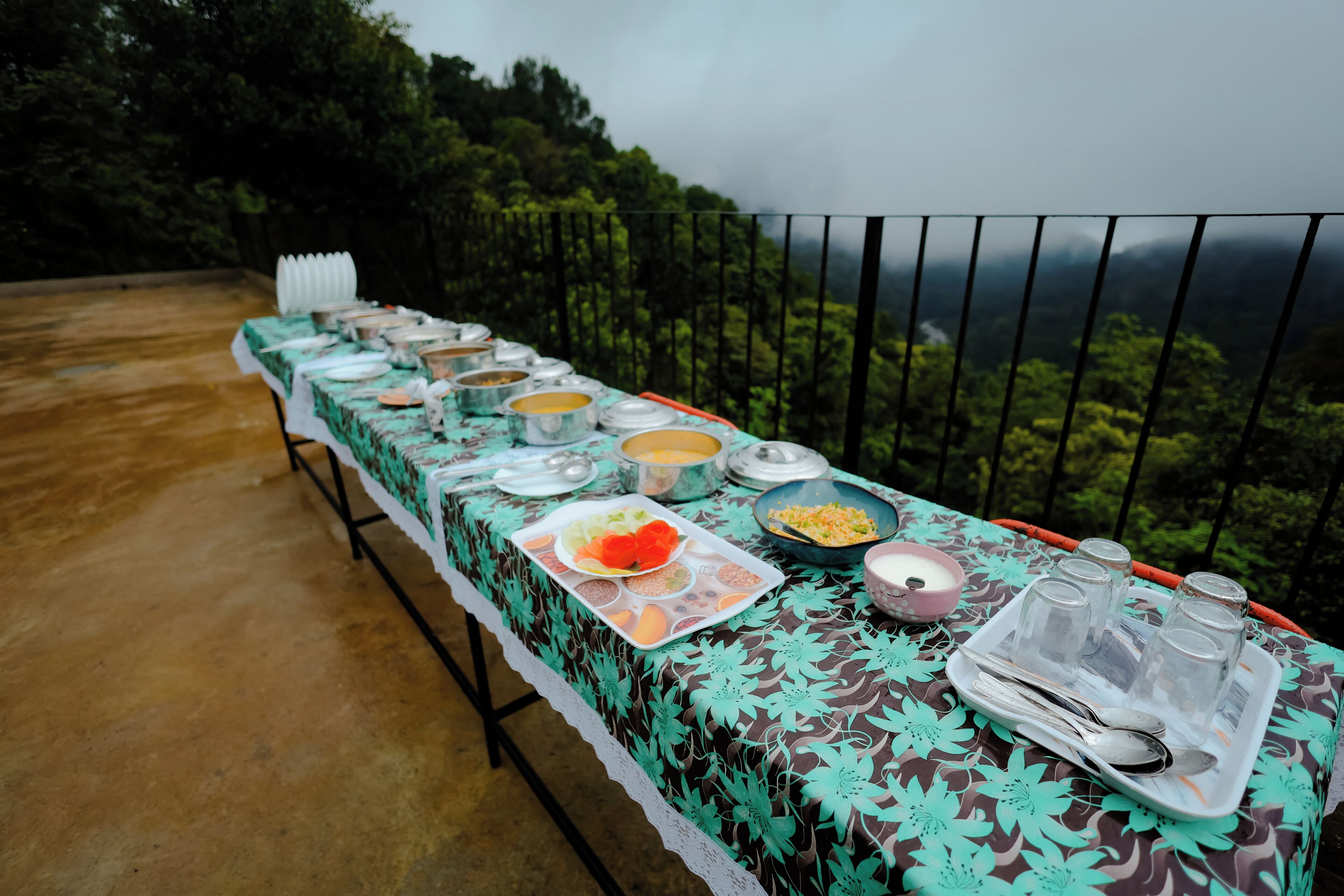 Mesa servida con diferentes platos y utensilios, rodeada por una barandilla con vista al bosque.