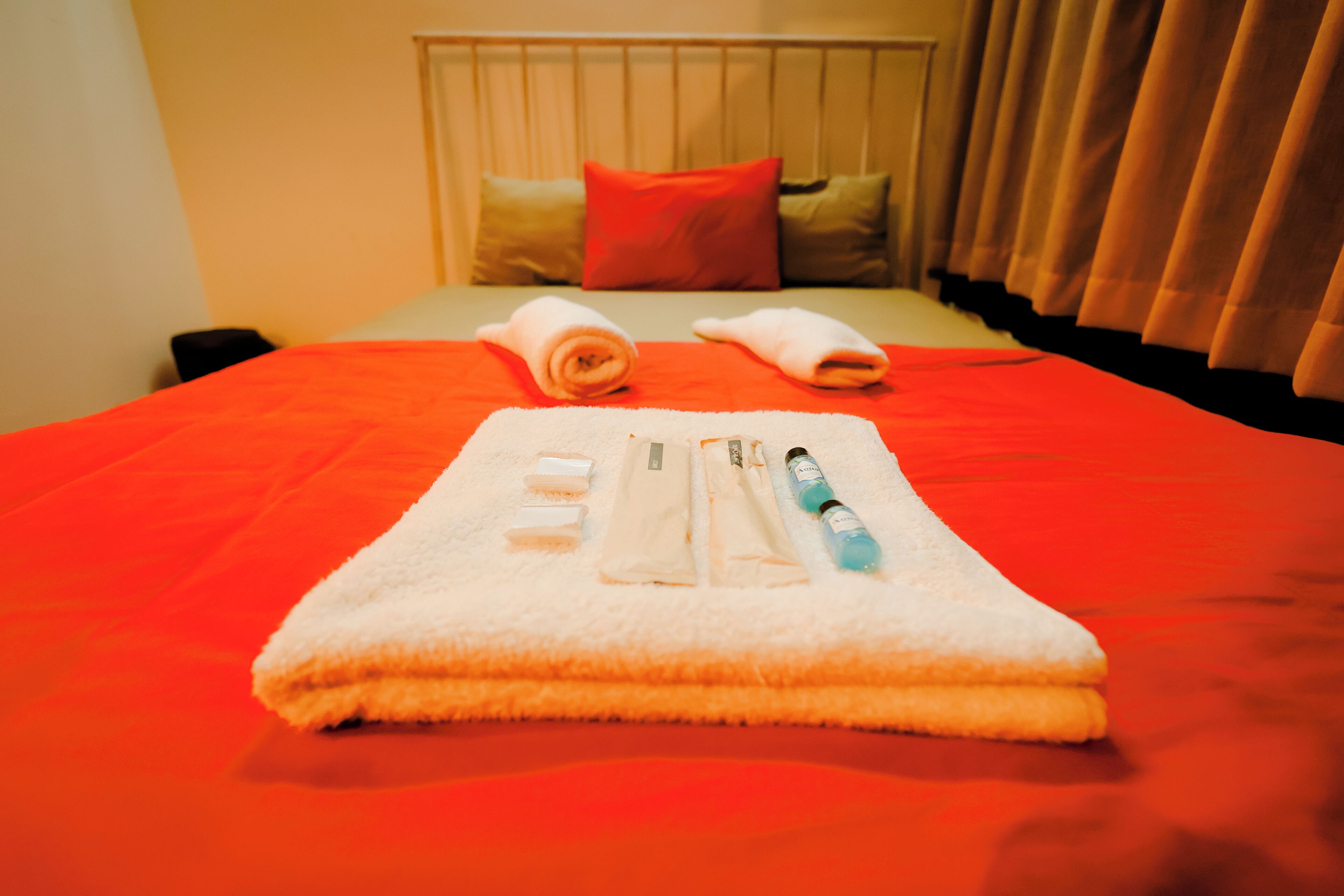 A neatly made bed with a red blanket, a white towel, and toiletries on a white towel.