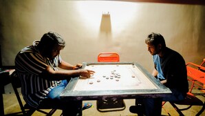 Two people playing a board game at a table with a simple, unadorned room in the background.