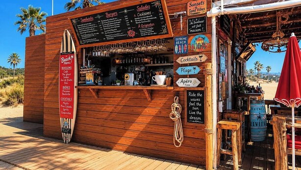 Een houten bar aan het strand met een menukaart, cocktails en uitzicht op het strand.