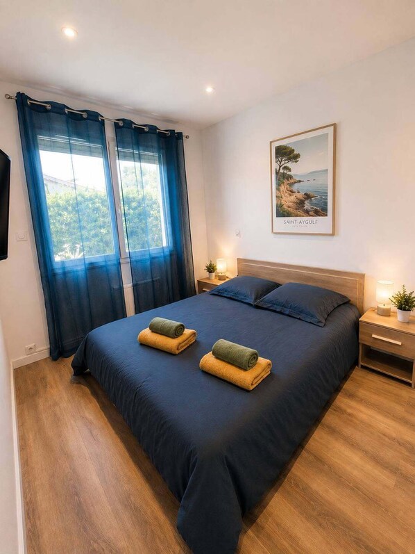 A bedroom with a bed, curtains, a nightstand, a lamp, a framed picture, and a wooden floor. - T3 air-conditioned with garden 900 m from the beach (Fréjus)