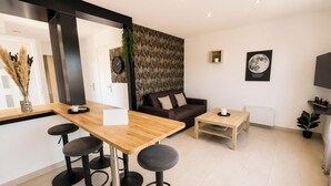 Een moderne woonkamer met een houten eettafel, twee zwarte barkrukken, een bruine bank, een klein houten bijzettafeltje en een klok aan de muur.