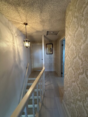 Um corredor com papel de parede estampado, uma porta branca, um lustre e uma escada com guarda-corpos brancos.