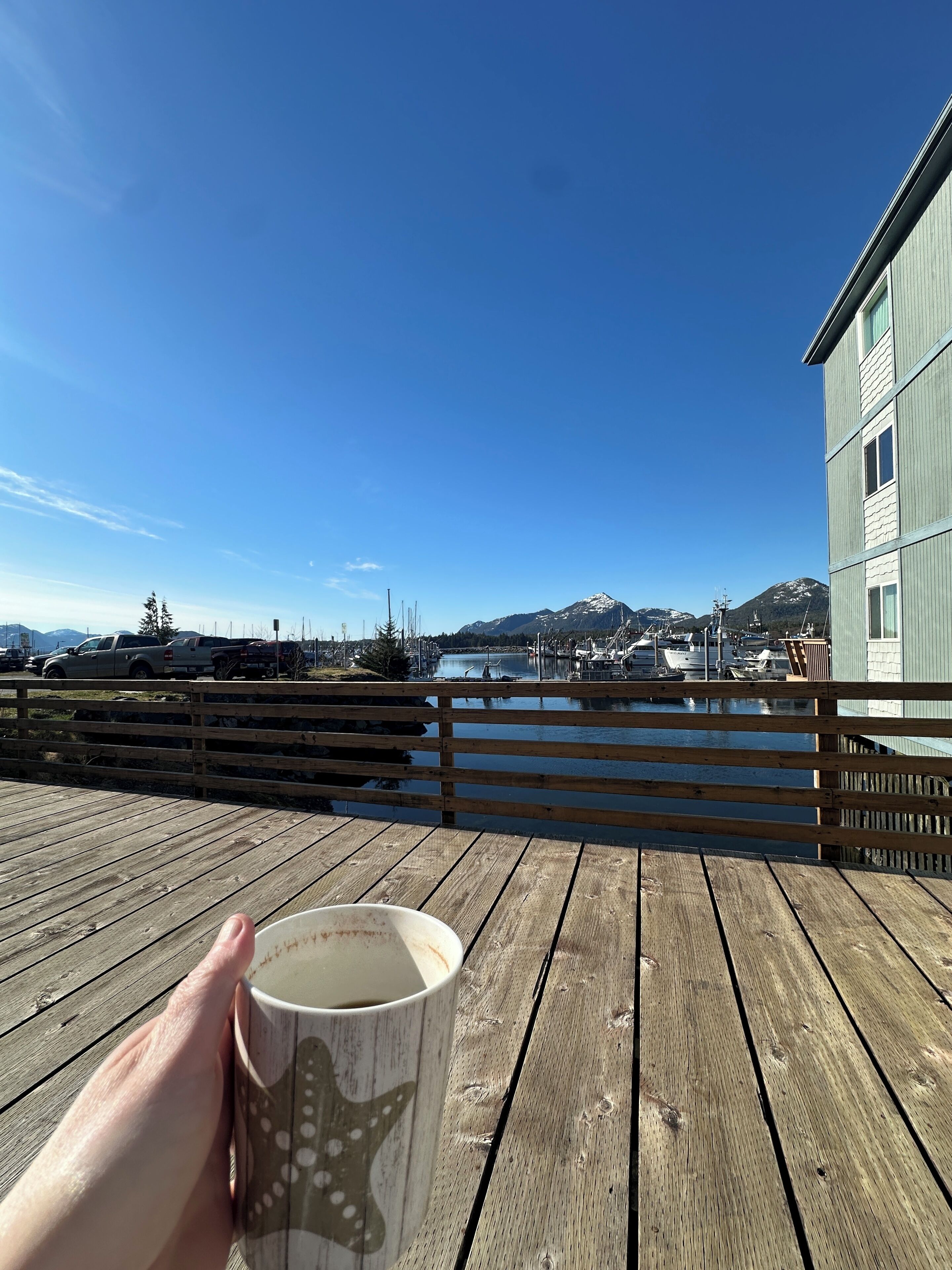 Una terraza de madera con vista a un puerto deportivo y montañas al fondo.