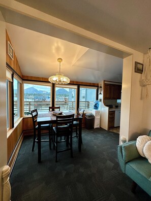A dining area with a table and chairs, a chandelier, a view of the cityscape, and a kitchenette in the background. - Oceanfront 1922 Charmer Meets Unbeatable Location In Ketchikan (Ketchikan)