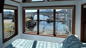 Ein Schlafzimmer mit großem Erkerfenster, einem Bett mit gemusterten Betttextilien und Blick auf einen Jachthafen und Berge.