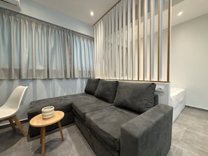 Uma sala de estar moderna com um sofá seccional cinza, uma mesa lateral redonda de madeira, uma cadeira branca e uma janela grande com cortinas.