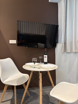 Quarto com televisão de tela plana na parede, uma mesa redonda pequena com duas cadeiras brancas, uma garrafa de vinho e dois copos de vinho.