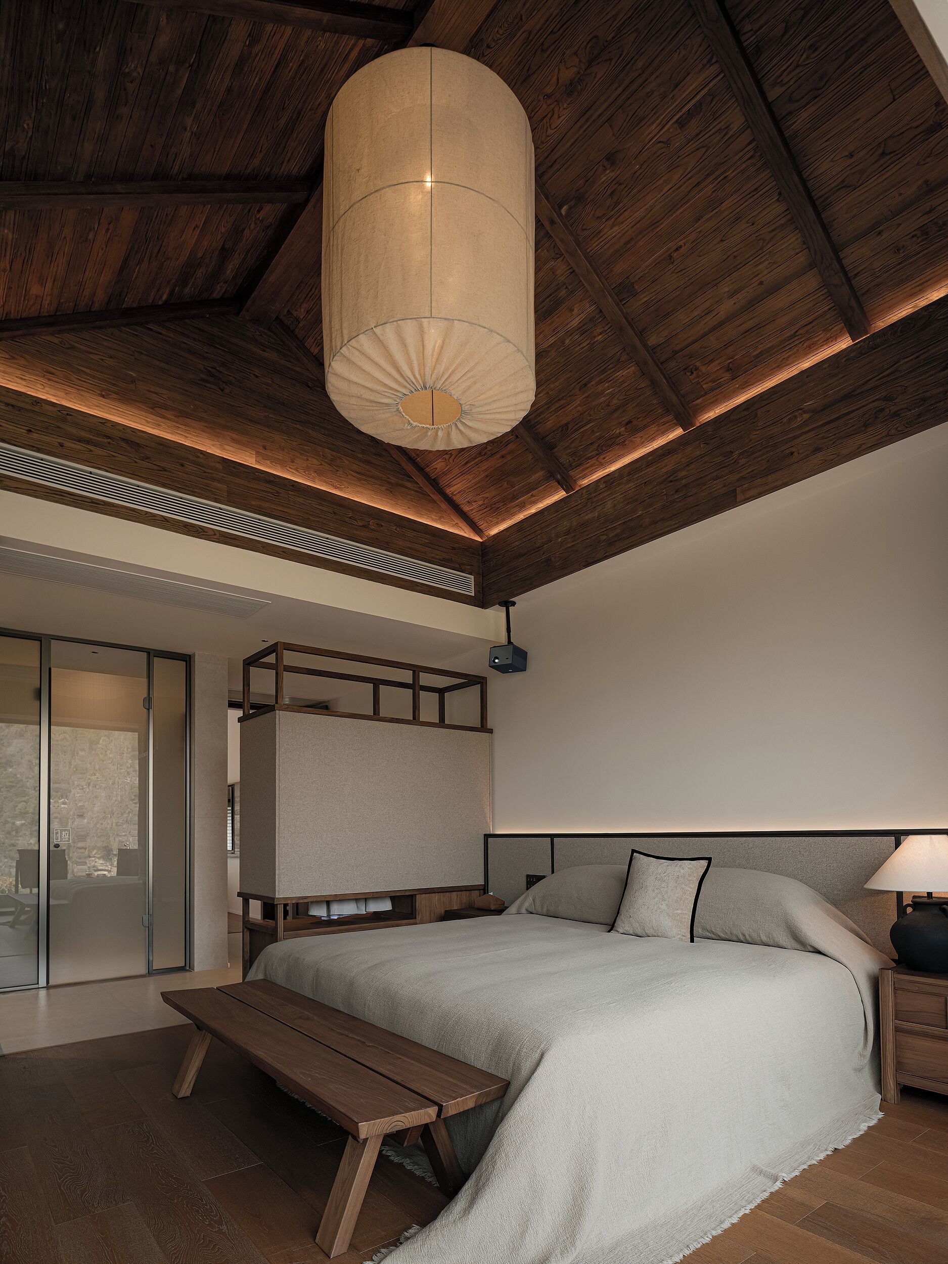 A bedroom with a bed, a bench, a hanging light fixture, and a view through a sliding door.