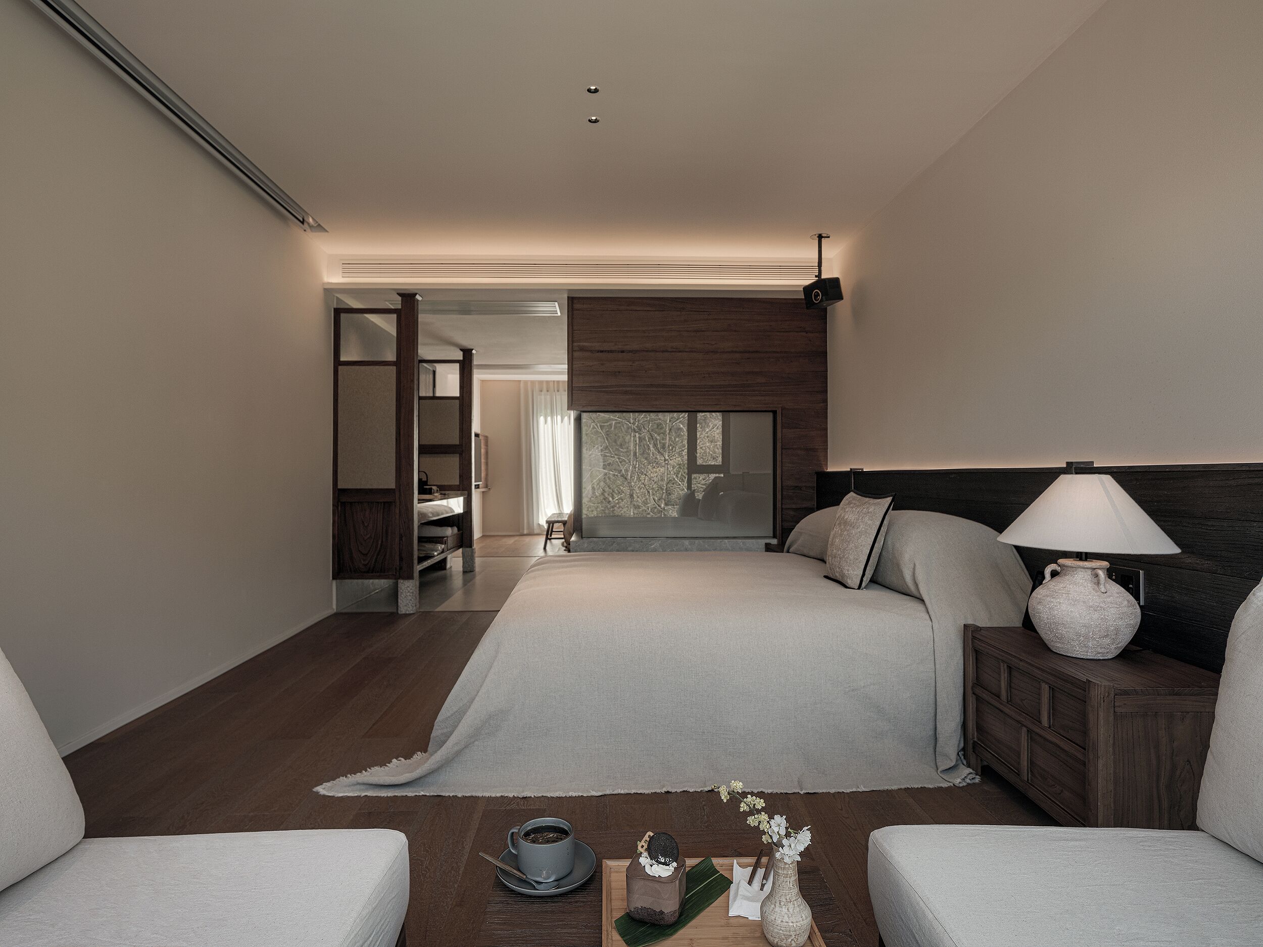 A modern bedroom with a large bed, bedside tables, a nightstand, and a view of a room with a window.
