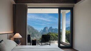A bedroom with a bed, a nightstand, a lamp, a chair, and a view of a mountainous landscape through a large glass door. - Zhangjiajie national park Oxgen Resort (Zhangjiajie)
