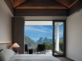 A bedroom with a bed, a nightstand, a lamp, a chair, and a view of a mountainous landscape through a large glass door.