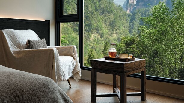 A room with a bed, a chair, and a table with a view of a forest and mountains. - Zhangjiajie national park Oxgen Resort (Zhangjiajie)