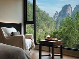 A room with a bed, a chair, and a table with a view of a forest and mountains.