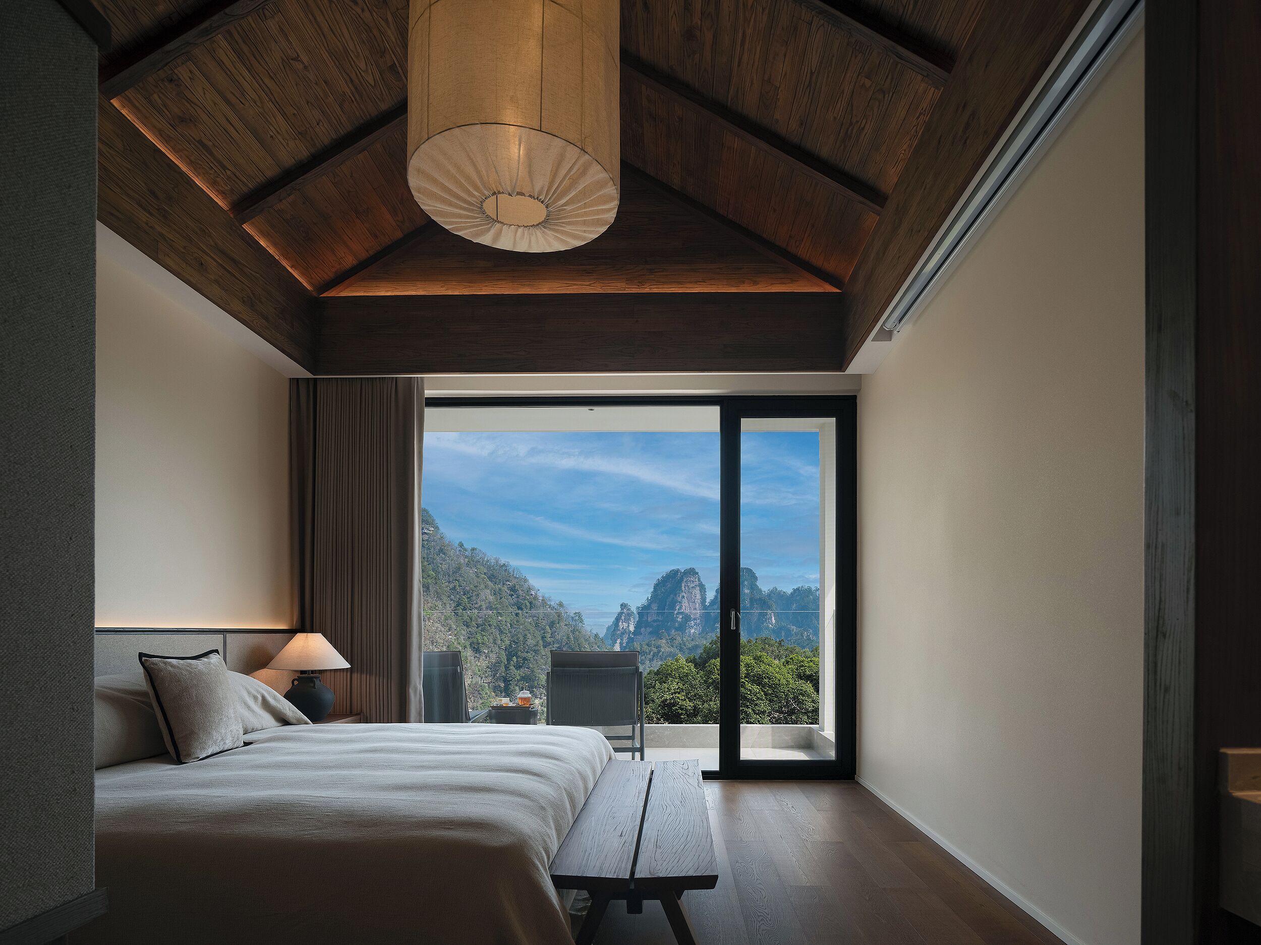 A bedroom with a bed, a nightstand, a lamp, a bench, and a large sliding glass door leading to a balcony with a mountain view.