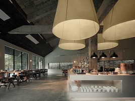 A spacious room with large windows, a long table with chairs, and a counter with kitchen appliances.