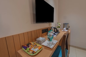 A hotel room with a wooden panel wall, a flat-screen TV mounted above a wooden counter, a coffee maker, a kettle, and a tray with snacks. - Hotel 7 Skye (Surat)
