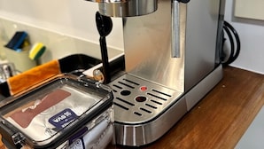 A stainless steel coffee machine with a digital pressure gauge, a water reservoir, and a clear plastic container with a label. - *Quiet, Cozy & Family-Friendly | Pool, Gym, Shops & Transport Nearby (Northshore)