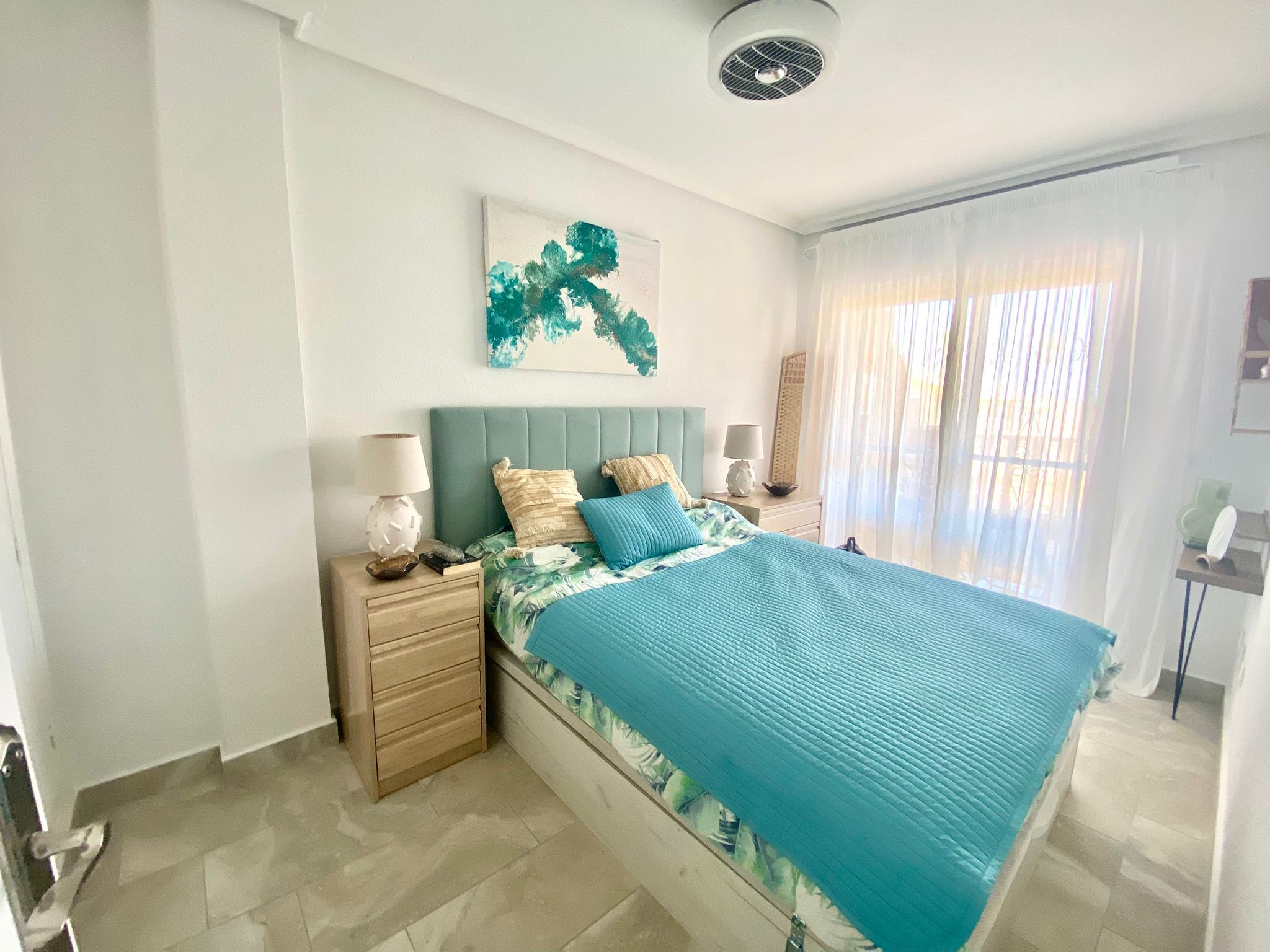 Un dormitorio con cama, mesitas de noche, una obra de arte colgada en la pared y una ventana con cortinas.