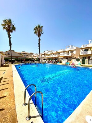 A swimming pool with a clear blue water, surrounded by palm trees and buildings with balconies. - Apartment in Cabo Roig, 300 meters from the beach (VC)
