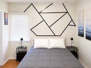 Un dormitorio con una cama, dos mesitas de noche, una ventana con persianas y un cuadro de pared con motivos geométricos.