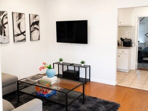 Un salon moderne avec un téléviseur à écran plat, des œuvres d’art abstraites, une table basse avec des livres et un vase, un canapé, et un coin cuisine à l’arrière-plan.